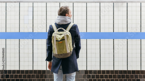 Lonely adult handsome man in a blue coat with a green backpack behind standing on the platform waiting for the train. Selective focus on male. Copy space. Morning commute. Urban life concept.