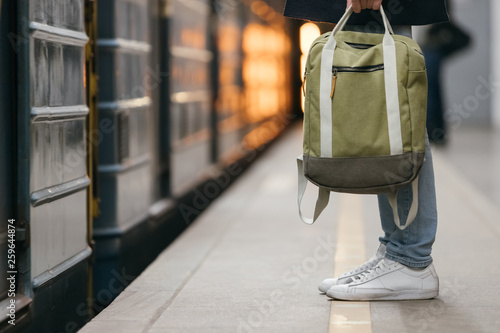 Close up photo of male waiting subway train on platform. Well-dressed handsome man in white sneakers and jeans with backpack in hand standing on station. Modern citizen of big city. Lifestyle concept.