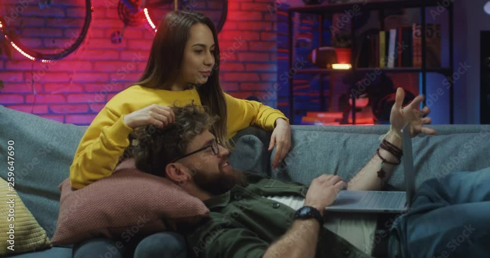 Attractive and happy Caucasian young girlfriend and boyfriend resting on the sofa with laptop computer at night, man lying on woman's laps while tapping and they looking on the screen. Attractive and happy Caucasian young girlfriend and boyfriend resting on the sofa with laptop computer at night, man lying on woman's laps while tapping and they looking on the screen.