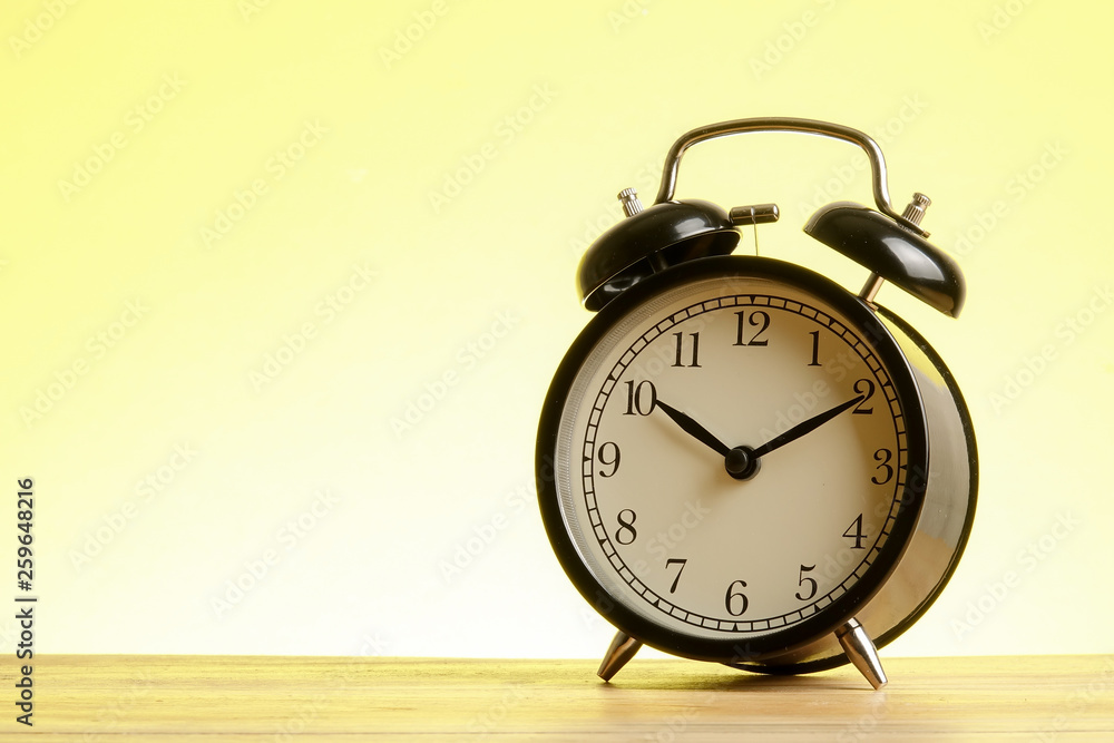 Black alarm clock on the wooden surface against the yellow background. Black alarm clock on the wooden surface against the yellow background.