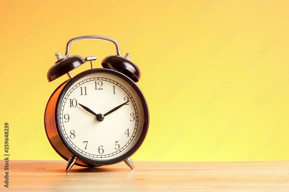 Black alarm clock on the wooden surface against the yellow background. Black alarm clock on the wooden surface against the yellow background.