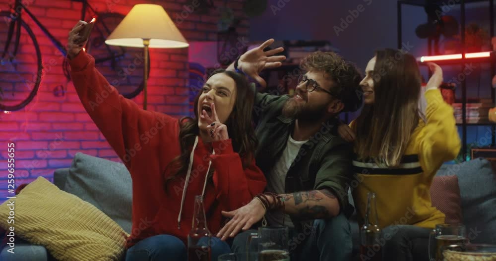 Young Caucasian stylish guy and two girls sitting on the couch in the living room at night and taking cheerful selfie photos while grimacing on the smartphone camera. Young Caucasian stylish guy and two girls sitting on the couch in the living room at night and taking cheerful selfie photos while grimacing on the smartphone camera.