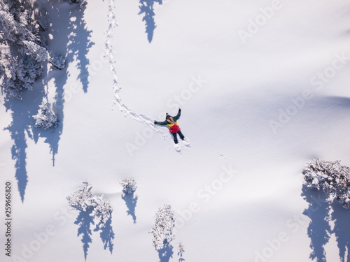 Winter aerial view portrait of Snowboarder female lying in clean snow between Christmas trees. Snowy mountain in ski resort, sunny winter holiday