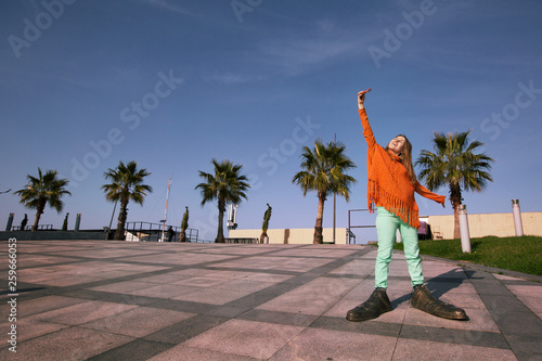 Young happy woman wearing orange poncho, bid funny shoes, doing selfie in city square with palm trees and embankment in Batumi, Georgia