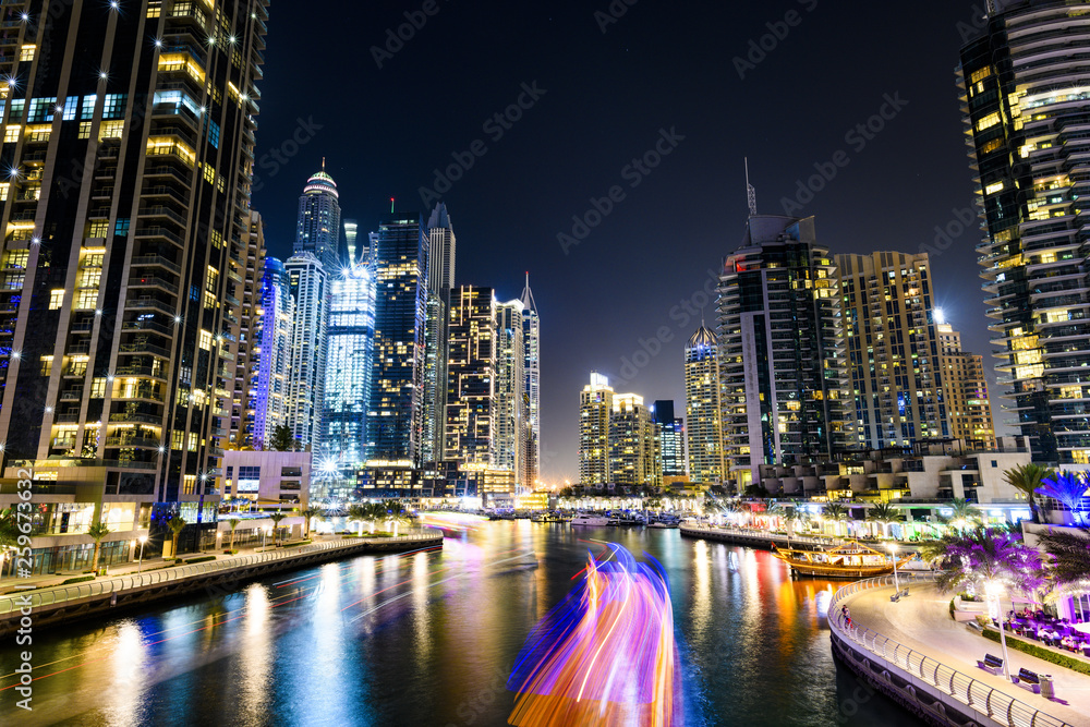 Stunning view of the Dubai Marina at dusk with illuminated skyscrapers in the background and a light trails left by a yacht sailing in the foreground. Dubai, United Arab, Emirates.