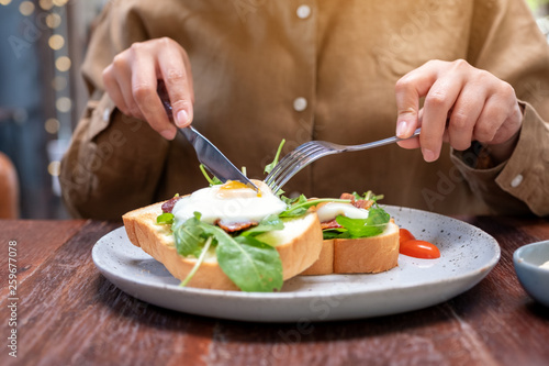 A woman eating breakfast sandwich with eggs, bacon and sour cream by knife and spoon in a plate on wooden table
