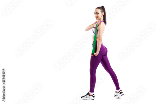 A young woman coach in a sporty purple short top and gym leggings exercise for your hands with sport fitness rubber bands on a white isolated background in studio