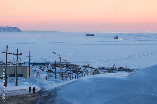 View of the bay, covered with ice. In the distance among the ice floes two cargo ships. On the road to the sea are two people. Big snowdrifts. Magadan, Nagaev Bay, Sea of ​​Okhotsk, Siberia, Russia. © Andrei Stepanov