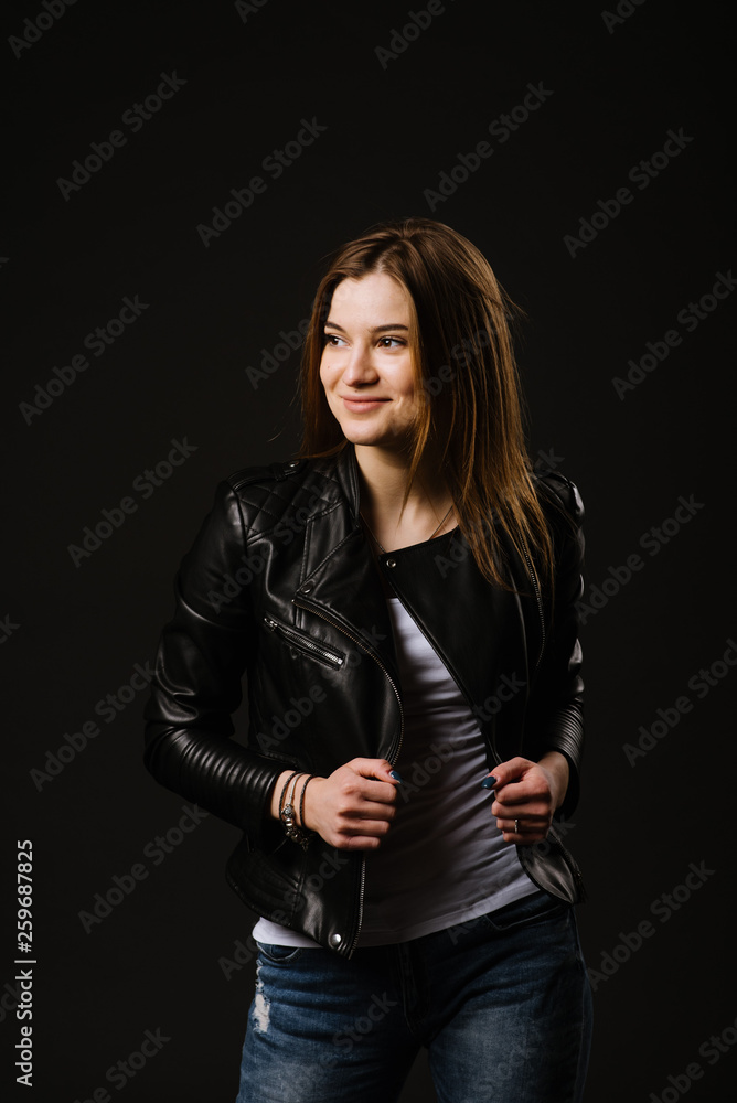 Young caucasian woman wearing a leather jacket and jeans posing on the black background Young caucasian woman wearing a leather jacket and jeans posing on the black background