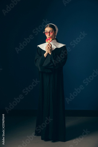 Waiting for new chances or answers. Medieval young woman as a nun in vintage clothing, white mutch and red sunglasses eating sweets and standing on dark blue background. Concept of comparison of eras. photo