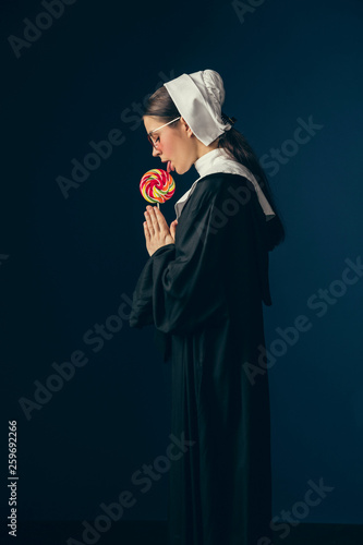 Forgive for passion for sweet. Medieval young woman as a nun in vintage clothing, white mutch and red sunglasses eating sweets and standing on dark blue background. Concept of comparison of eras. photo