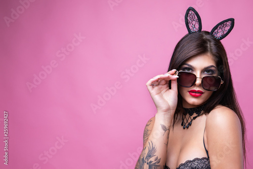Cute sexy brunette with hooked ears of hare and sunglasses poses on pink background