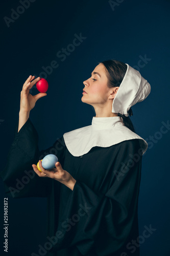 Find the best at last. Medieval young woman as a nun in vintage clothing and white mutch standing with the Easter colored red, yellow eggs and on dark blue background. Concept of comparison of eras. photo