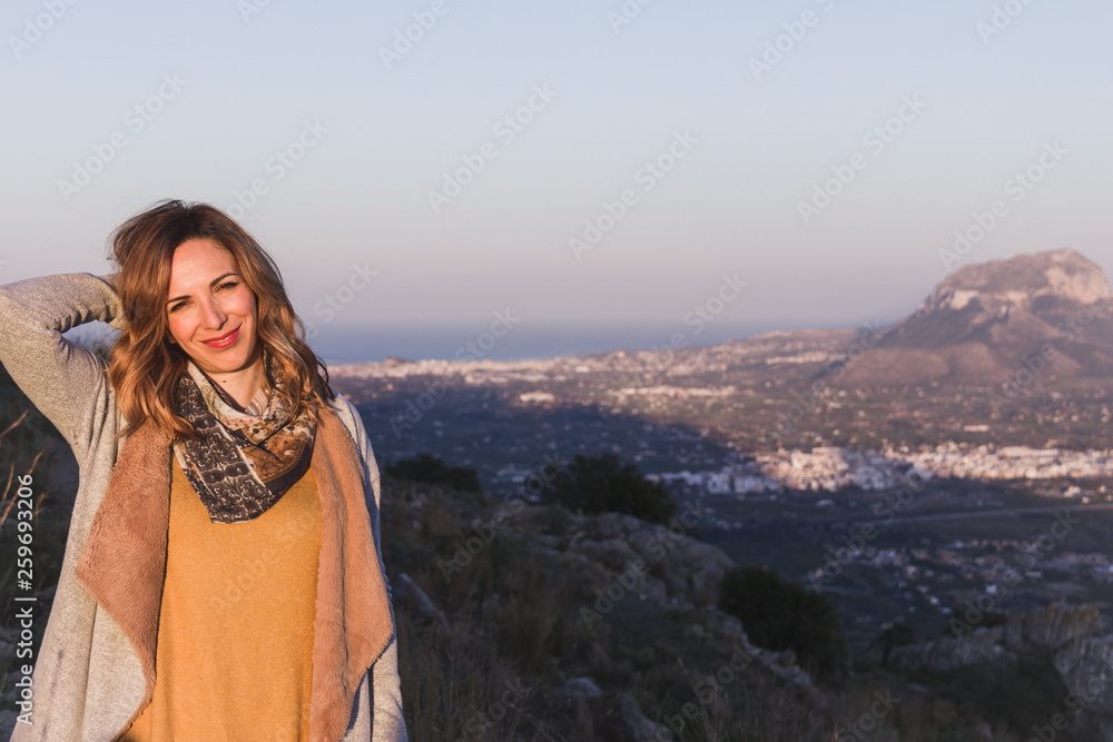 Naklejka premium Portrait of a young woman with ochre colored clothes, posing in a beautiful landscape at sunset. Montgo mountain is in the background, in Spain