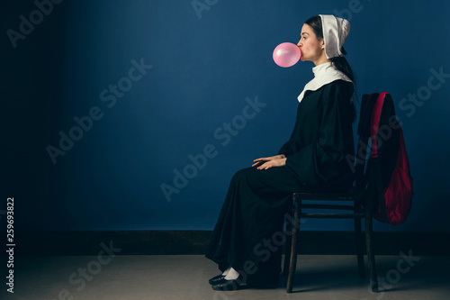 New emotions of gray days. Medieval young woman as a nun in vintage clothing, white mutch and red bag sitting on the chair on blue background. Blowing bubble of gum. Concept of comparison of eras. photo