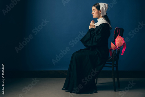 Memories of youth or crazy weekend. Medieval young woman as a nun in vintage clothing and white mutch sitting on the chair with bright pink bra on blue background. Concept of comparison of eras. photo