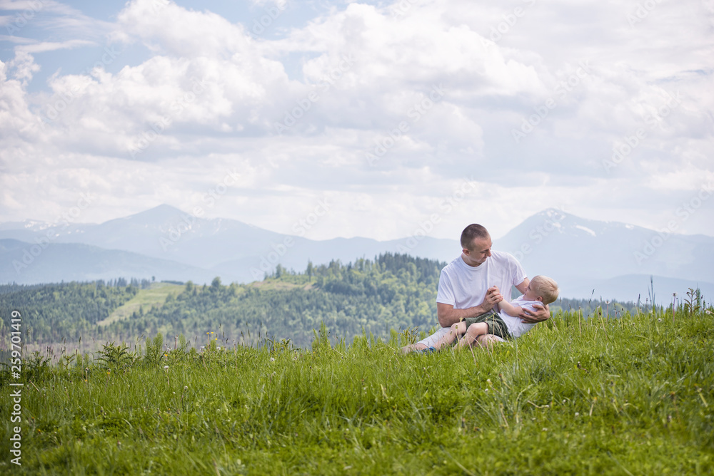 Happy father with his little son on his knees sits against a background of green forest, mountains and sky with clouds. Friendship concept