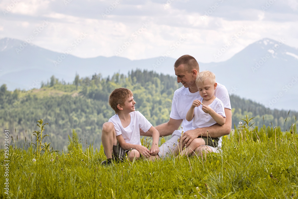Fototapeta premium Happy father with his two young sons sitting on the grass on a background of green forest, mountains and sky with clouds. Friendship concept.