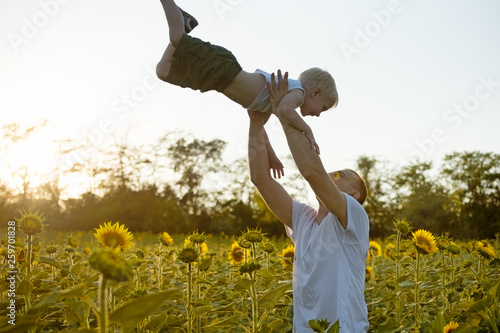 Father throws up his little son on sunflowers field against the sky