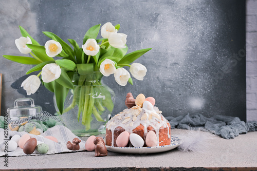Bouquet of white tulips in a vase on a gray background. Shokloy eggs of different colors and Easter cake with cream, as a symbol of the Easter holiday. Spring card with flowers and sweets. Copy space.