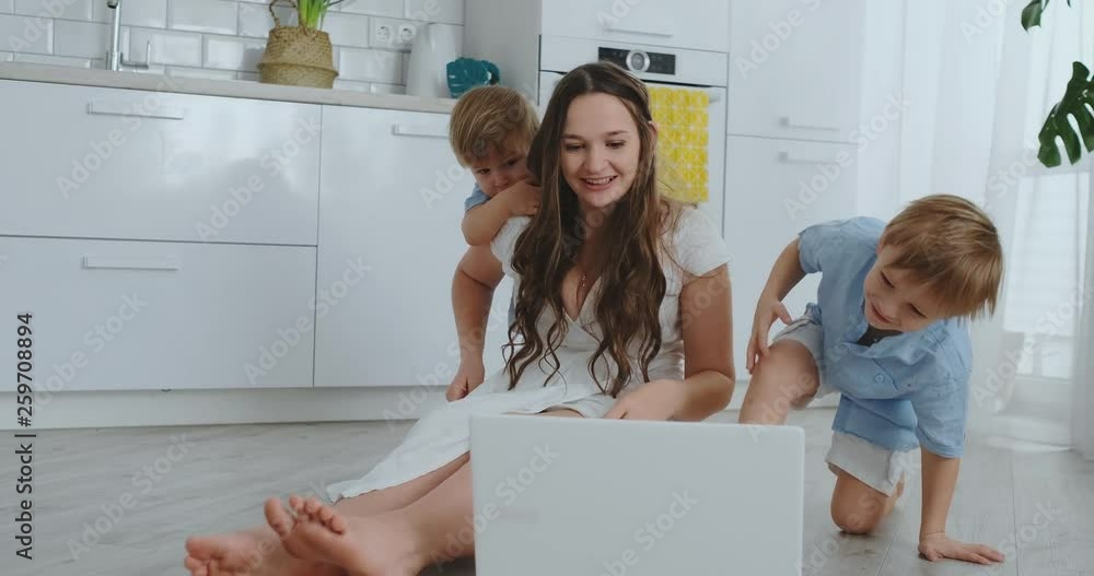 Modern technology. Modern apartment loving mom and two small sons sitting on the floor in the living room look at the laptop screen. Children with mom play on a laptop. Modern technology. Modern apartment loving mom and two small sons sitting on the floor in the living room look at the laptop screen. Children with mom play on a laptop.