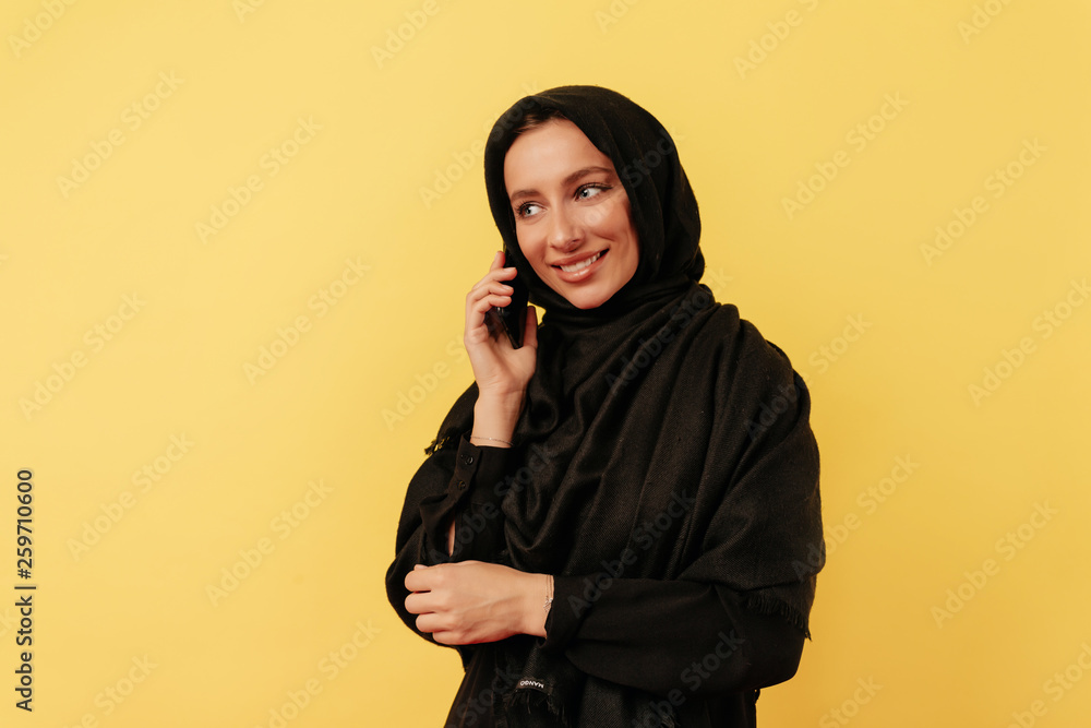 Eastern woman wearing burqa using gadget and talking on smartphone over isolated yellow background Eastern woman wearing burqa using gadget and talking on smartphone over isolated yellow background