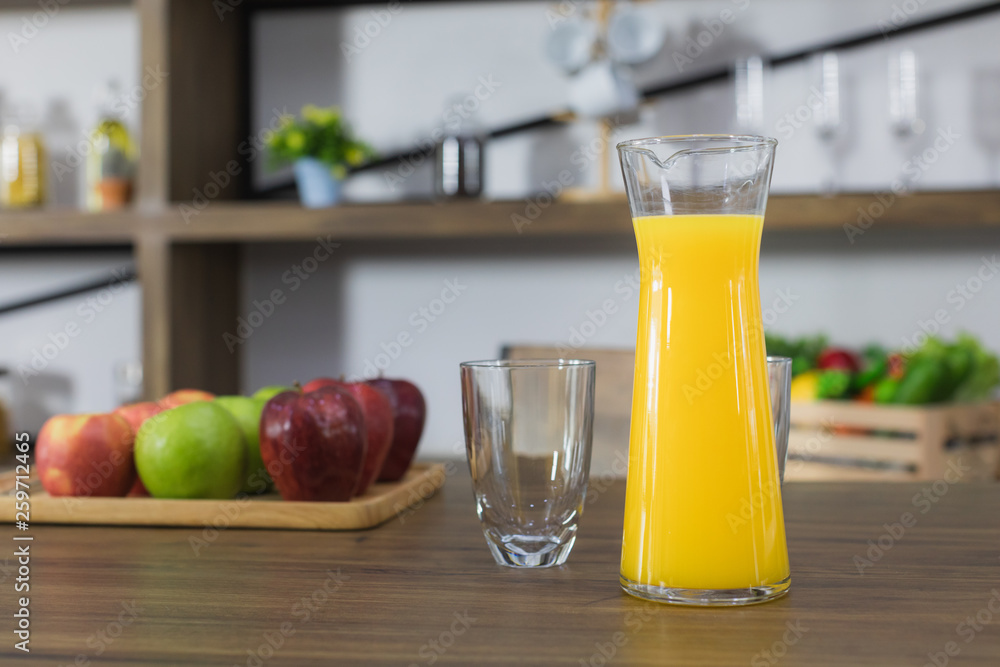 Close up image of a jug of freshly squeezed orange juice and drinking glass on the dark wooden table top, with a blur green and red apple and kitchen room background Close up image of a jug of freshly squeezed orange juice and drinking glass on the dark wooden table top, with a blur green and red apple and kitchen room background