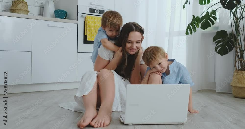 Modern apartment loving mom and two small sons sitting on the floor in the living room look at the laptop screen. Children with mom play on a laptop Modern apartment loving mom and two small sons sitting on the floor in the living room look at the laptop screen. Children with mom play on a laptop