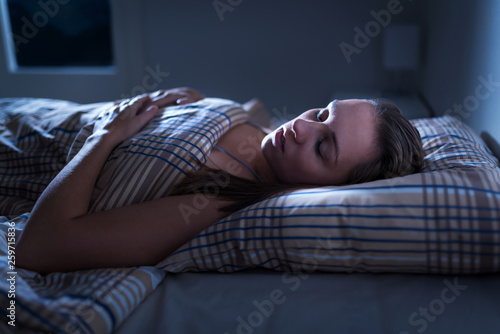 Calm and peaceful woman sleeping in bed in dark bedroom. Lady asleep at home in the middle of the night. Pillow, blanket and moonlight. Nightmare or sleep apnea concept.