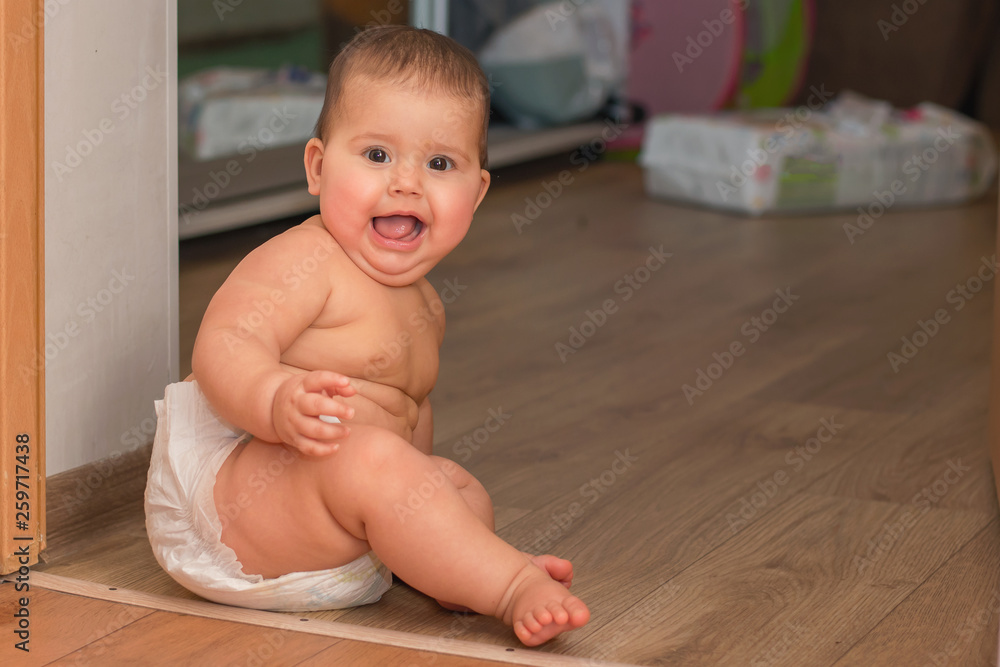 Joyful toddler girl in a diaper sitting on the floor and looking at the camera, lifestyle, close-up Joyful toddler girl in a diaper sitting on the floor and looking at the camera, lifestyle, close-up