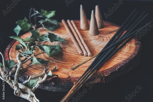 Examples of various types of incense - long black sticks, short hand made sticks and  small triangular cones. Black background, placed on a wooden plate cutting with ivy plant branch laying next to it photo