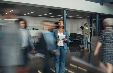 Beautiful black african business woman standing in a busy lobby of an office looking upwards, holding a tablet with people walking past in a blur