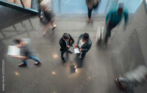 Two business people standing in the lobby of an office looking at a tablet while people are walking past in a blur photo