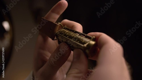 A man holds a copper cryptex with numbers. He calculates the cryptex code with his hand. At the end of the cryptex, a leather strap is put on the man’s finger. There is a dim light in room. Closeup. photo