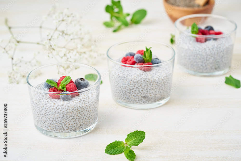 Chia pudding with fresh berries raspberries, blueberries. Three glass, light wooden background, side view, diagonal, flowers. Chia pudding with fresh berries raspberries, blueberries. Three glass, light wooden background, side view, diagonal, flowers.