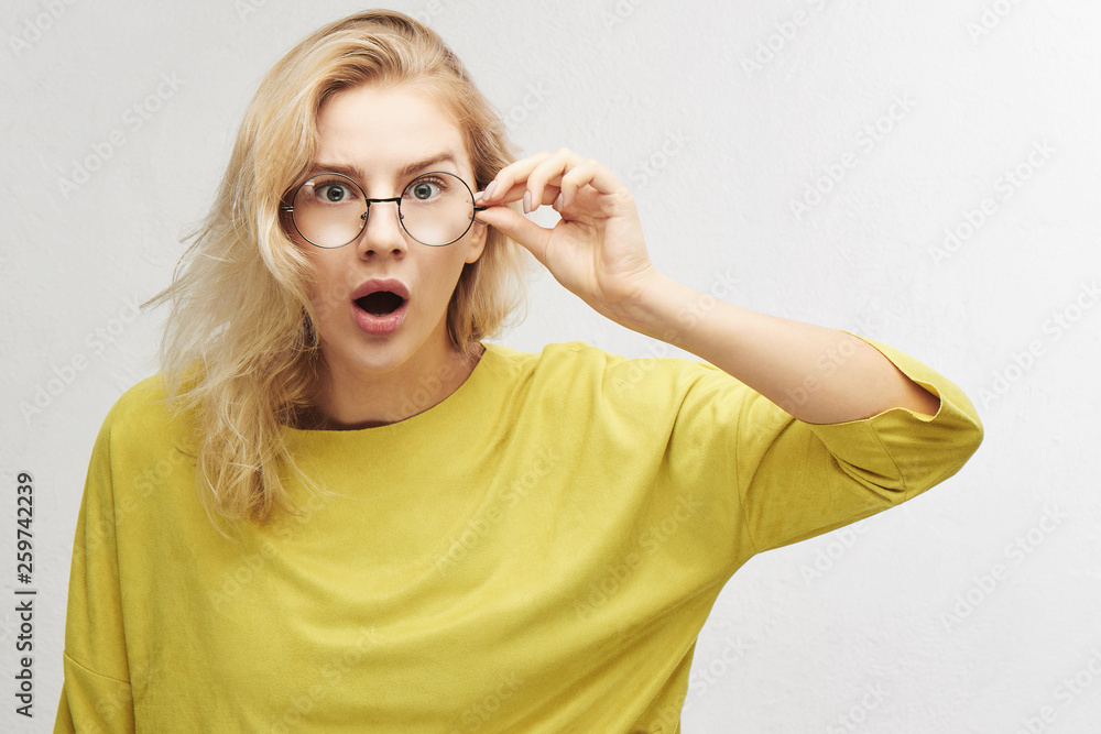 Surprised woman in round glasses with open mouth and bulging eyes looks into camera and sees something incredible and amazing isolated on white background - obrazy, fototapety, plakaty 