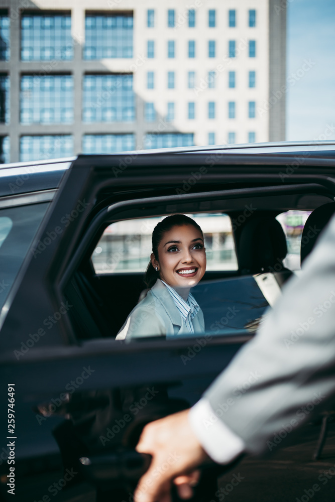 Good looking young business woman sitting on backseat in luxury car. She smiling and looking out. Transportation in corporate business concept.