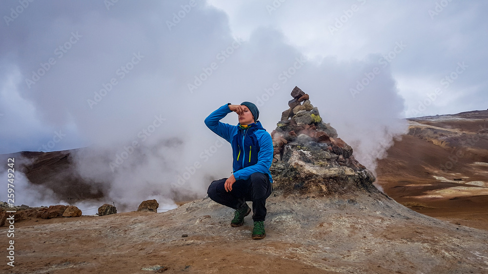 Young man wearing a blue squatting in front of a steaming fumaroles emitting sulfuric gas and pinching his nose to prevent from bad smell. Power of planet Earth.