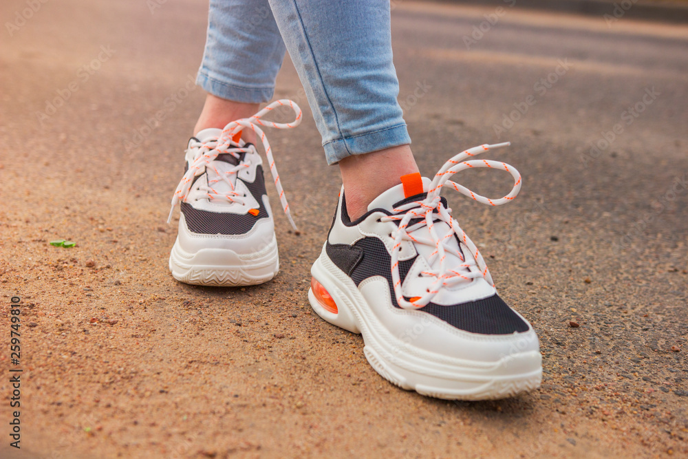 Girl in sneakers and jeans. Several pairs of sports shoes and legs. Stylish fashionable white women's leather sneakers on asphalt on a sunny day. Female legs with sneakers