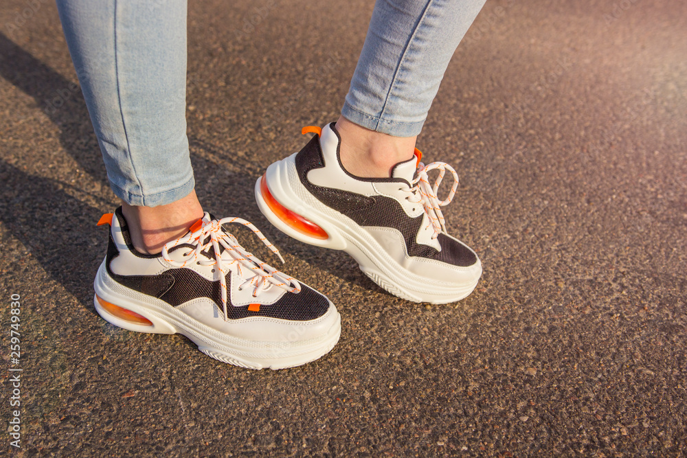 Girl in sneakers and jeans. Several pairs of sports shoes and legs. Stylish fashionable white women's leather sneakers on asphalt on a sunny day. Female legs with sneakers