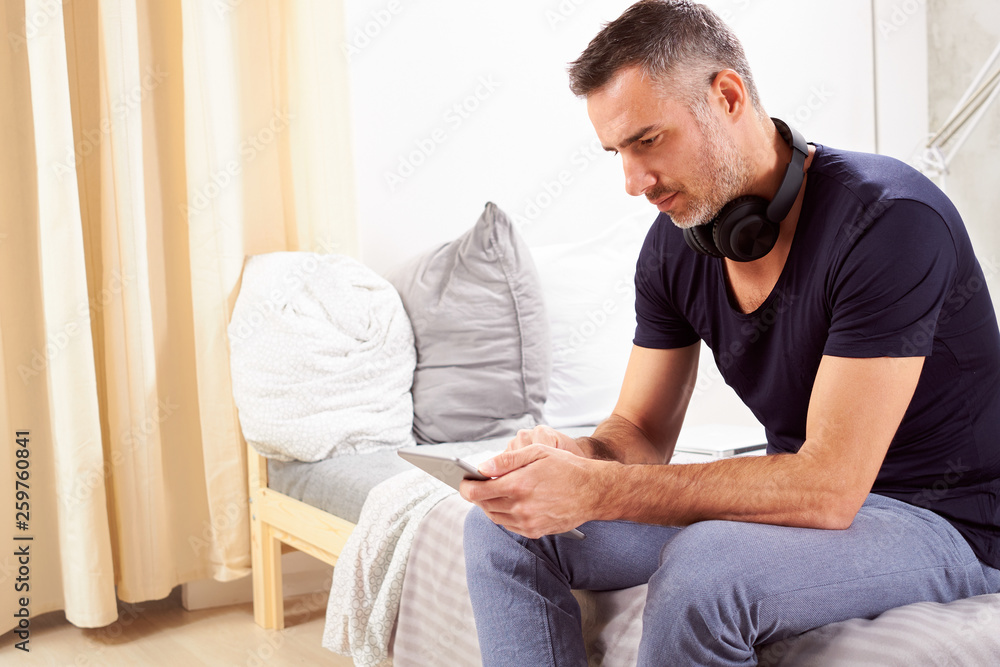 Caucasian man in forties sitting on bed in bedroom and using tablet.