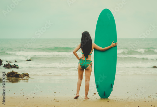 Sexy woman in green bikini standing with surfboard on beach