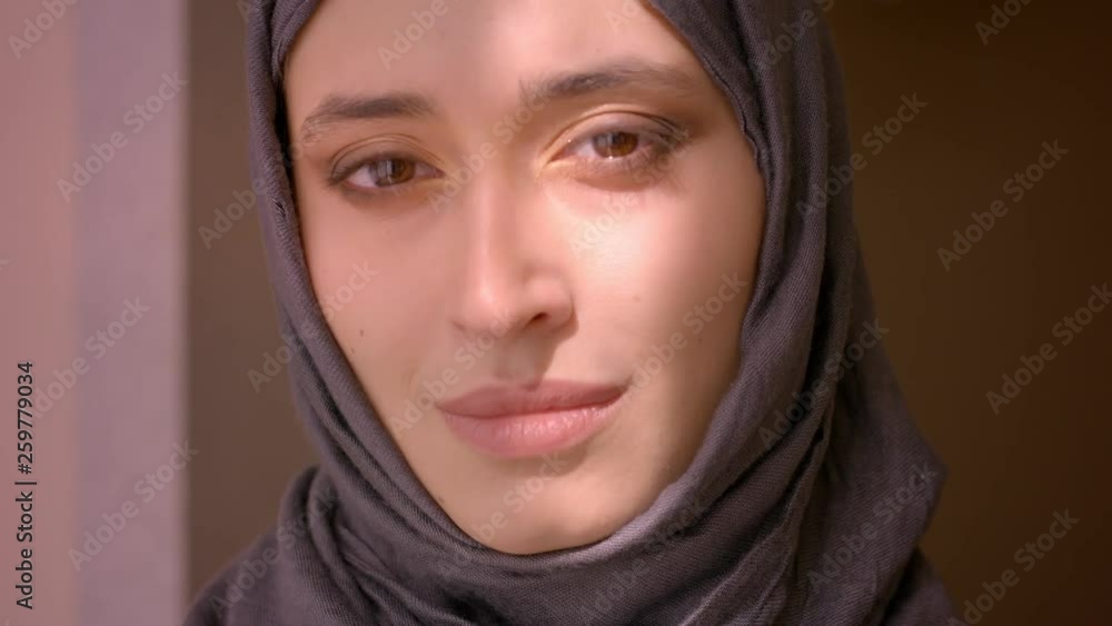 Closeup shoot of young attractive muslim female looking at camera and squinting her eyes indoors at home with sun shining on her face Closeup shoot of young attractive muslim female looking at camera and squinting her eyes indoors at home with sun shining on her face