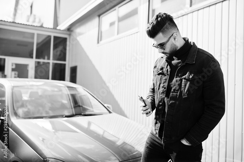 Fashion Arab man wear on black jeans jacket and sunglasses posed against business modern car. Stylish, succesful and fashionable arabian model guy looking at mobile phone. photo