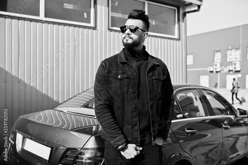 Fashion Arab man wear on black jeans jacket and sunglasses posed against business modern car. Stylish, succesful and fashionable arabian model guy.