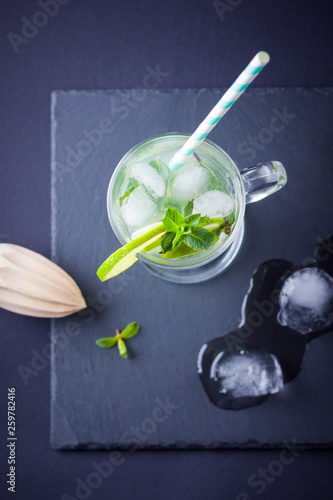 Mojito cocktail and ice chunks on black background. Mojito with lime and fresh mint on slate board. Summer refreshing cocktail and wooden juicer on dark background. Infused water
