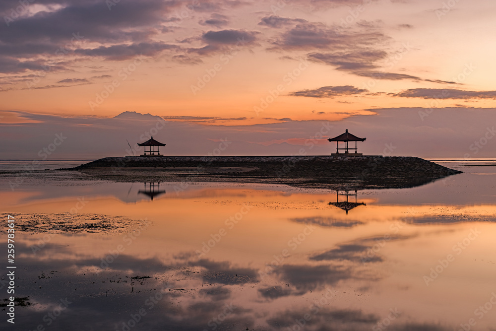Bali Sanur at sunrise on the beach. Beautiful calm water with 2 temples on a breakwater. Great reflection in the water with backlighting and great clouds