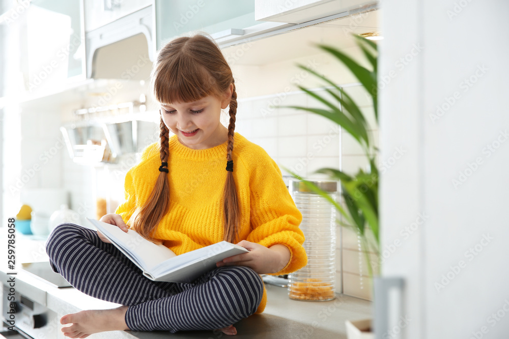 Cute little girl reading book in kitchen at home Cute little girl reading book in kitchen at home