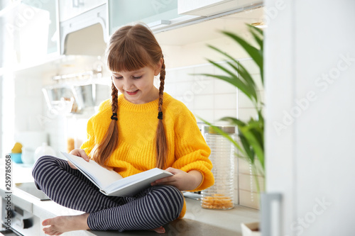 Cute little girl reading book in kitchen at home