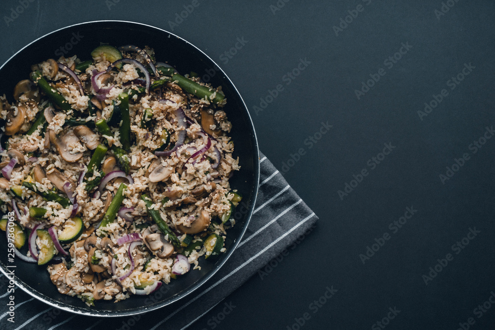 Brown rice with asparagus, courgette, mushrooms and red onion in a pan on stripy grey tea towel on black background with space of copy or text.